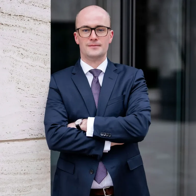 Rechtsanwalt Griemert Rechtsanwalt Raphael Griemert in Anzug mit Brille, siegesgewiss vor modernem Gebäude.
